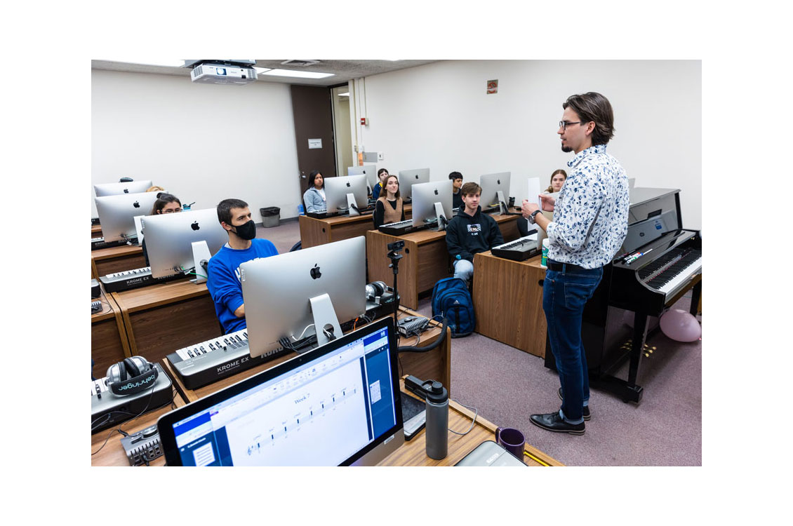 A professor stands in front of a classroom full of students sitting at computes with piano keyboards