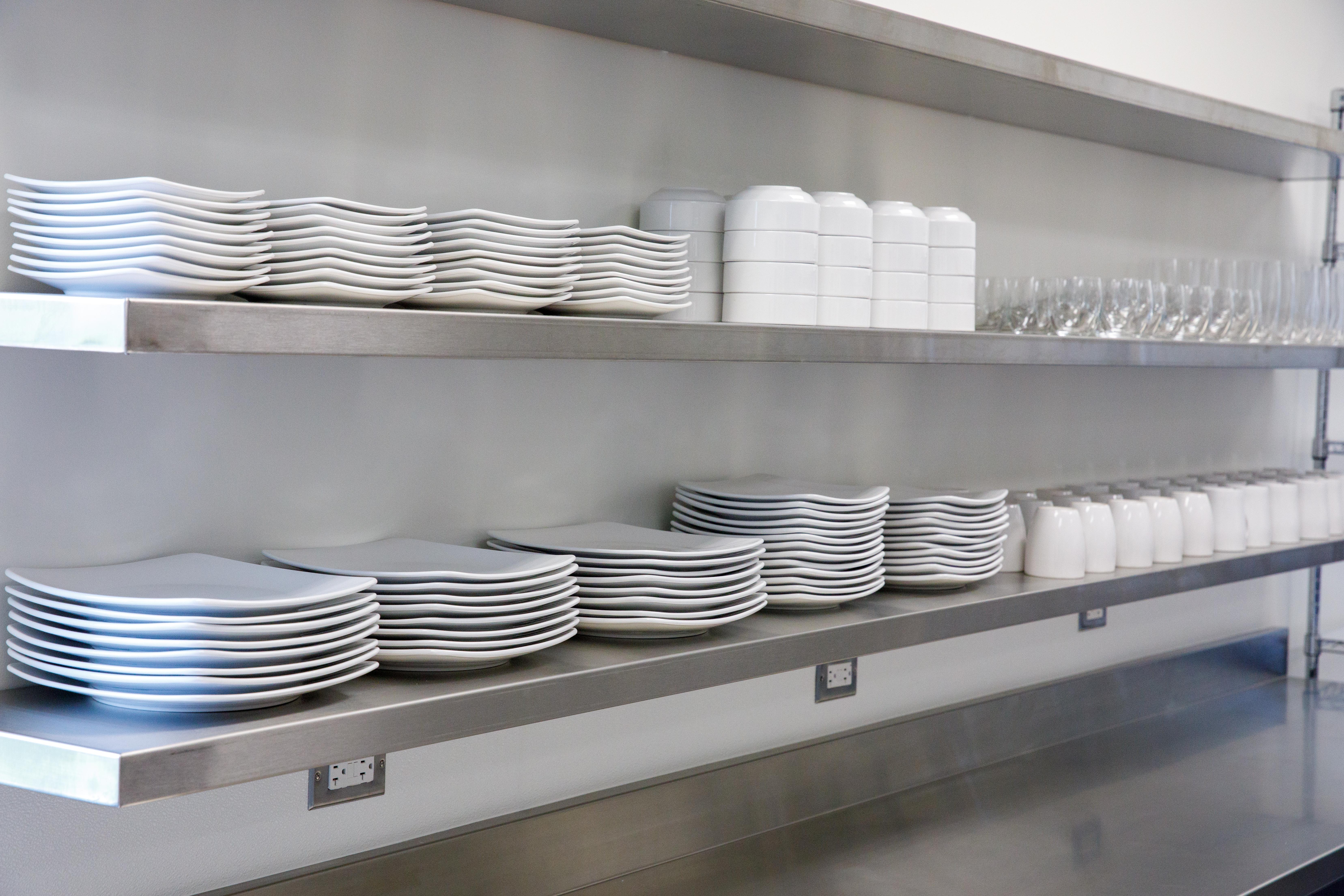 Close up of dozens of clean plates, bowls, cups and glasses neatly stacked on stainless steel shelves.