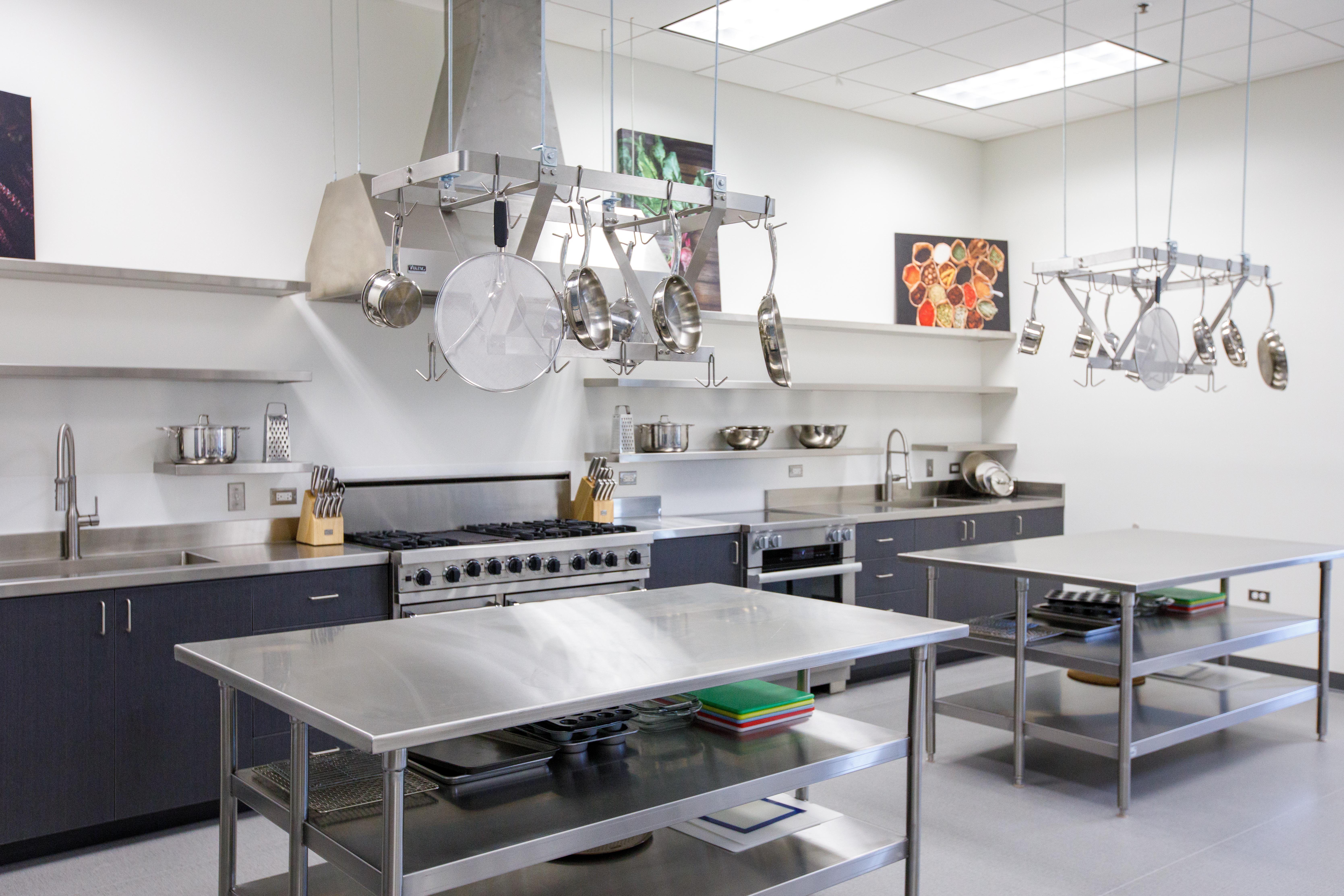 From another view, a clean, modern stainless kitchen area, with stainless steel prep tables and counters, pots and pans, a stove, sink