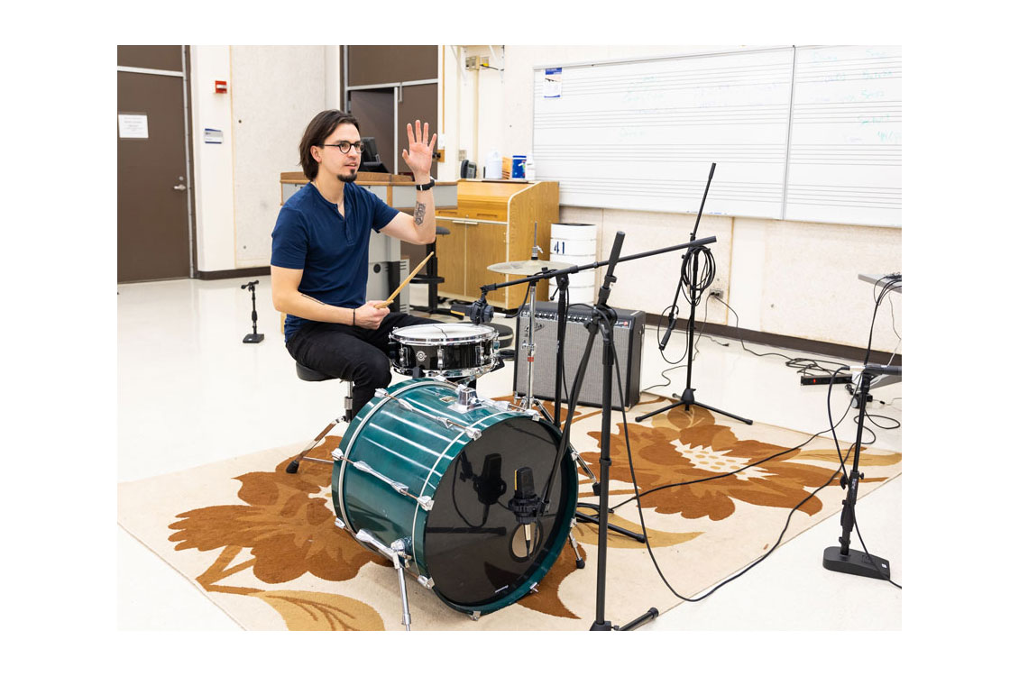 a professor sits behind a drum kit with his hand raised